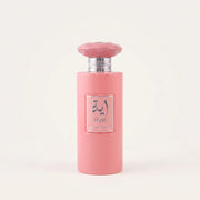 AYA Eau de Parfum cylindrical opaque pastel pink bottle topped with a matching faceted gem-shaped cap and textured silver collar, featuring a square silver-framed label with metallic silver Arabic calligraphy.