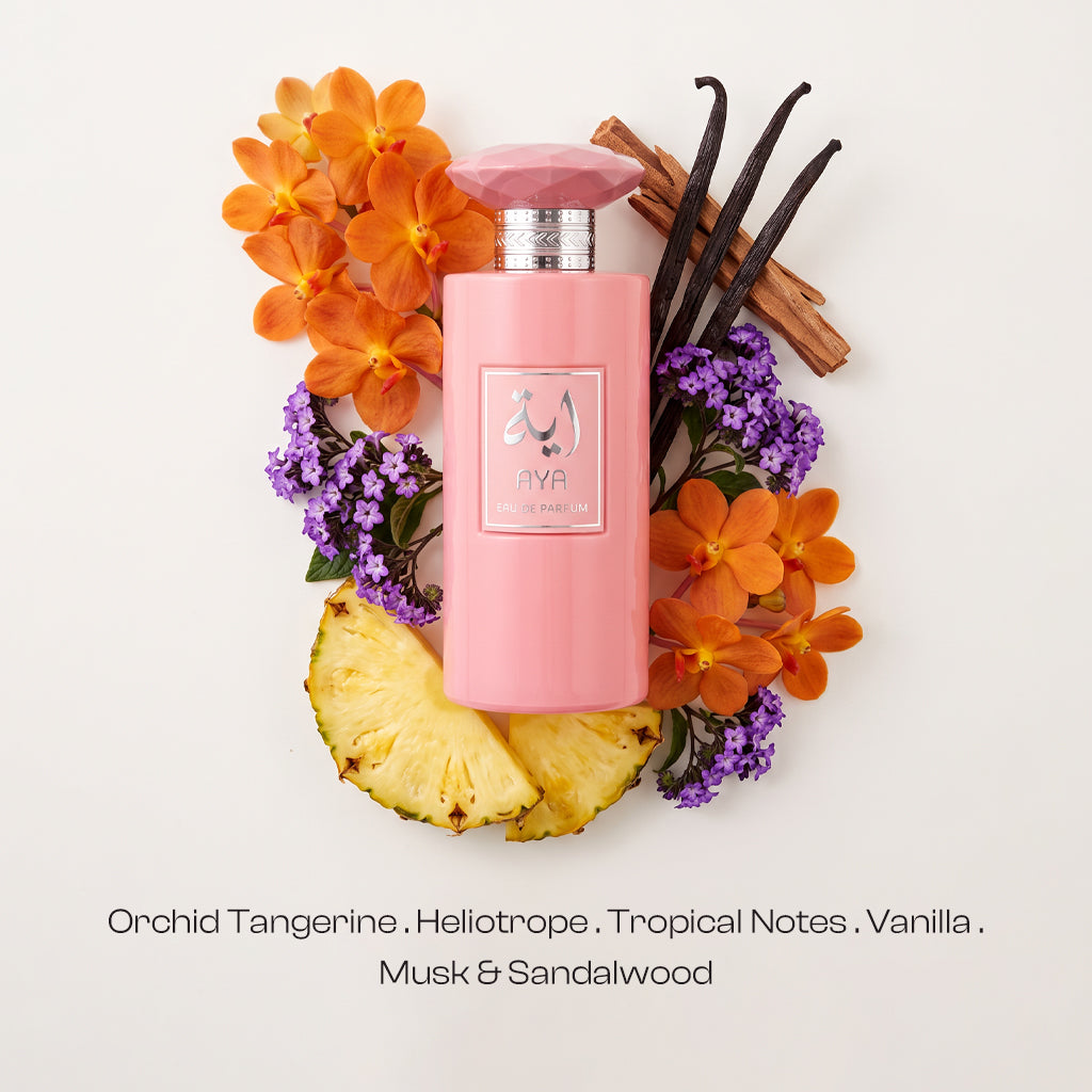 AYA Eau de Parfum pink bottle with silver accents arranged in a flat lay surrounded by orange orchids, purple heliotrope blossoms, fresh pineapple slices, vanilla pods, and cinnamon sticks on a off-white background with fragrance notes below.