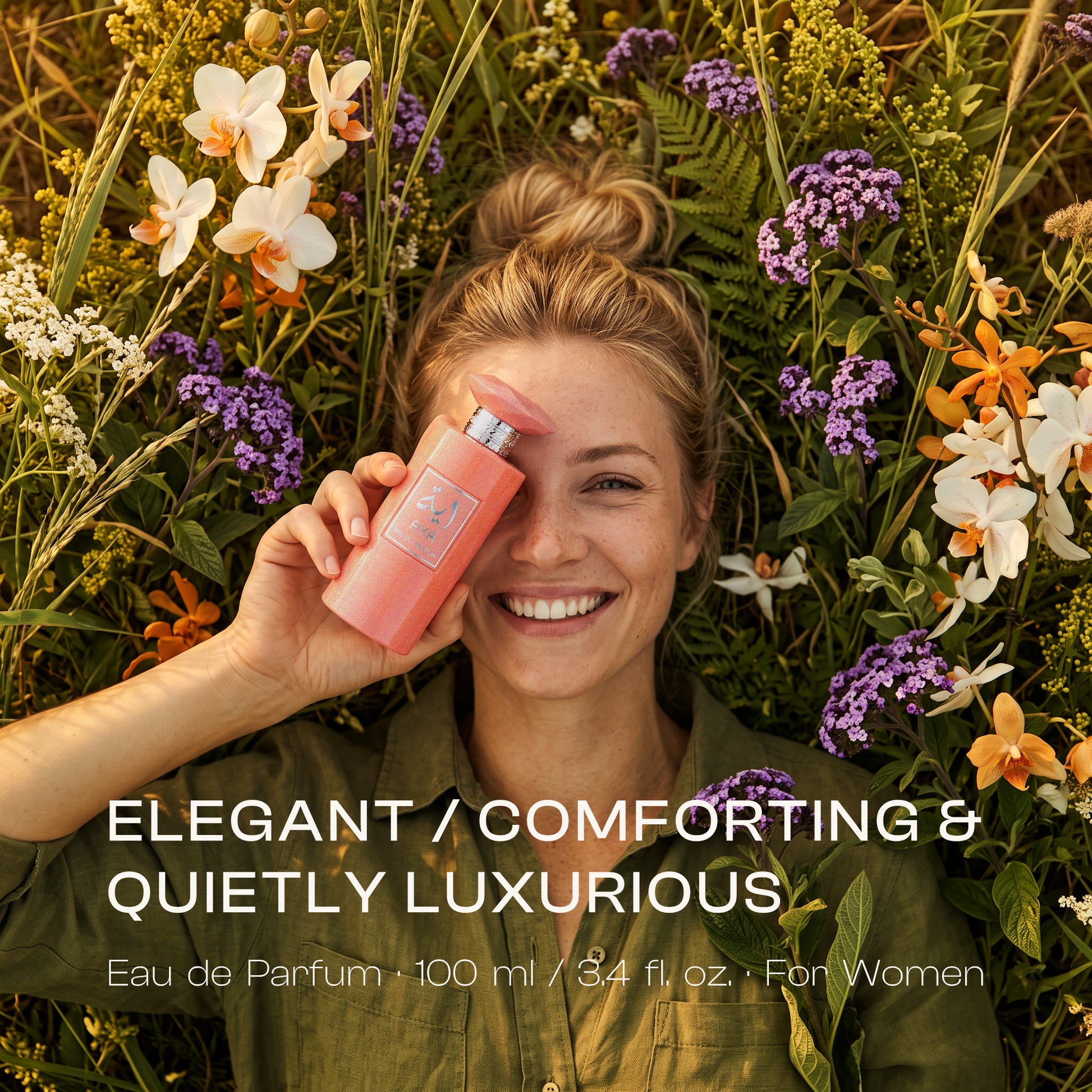 Smiling blonde woman lying in a field surrounded by white, orange, and purple flowers, holding a shimmery coral pink AYA Eau de Parfum bottle over one eye, with text overlay reading "ELEGANT / COMFORTING & QUIETLY LUXURIOUS".