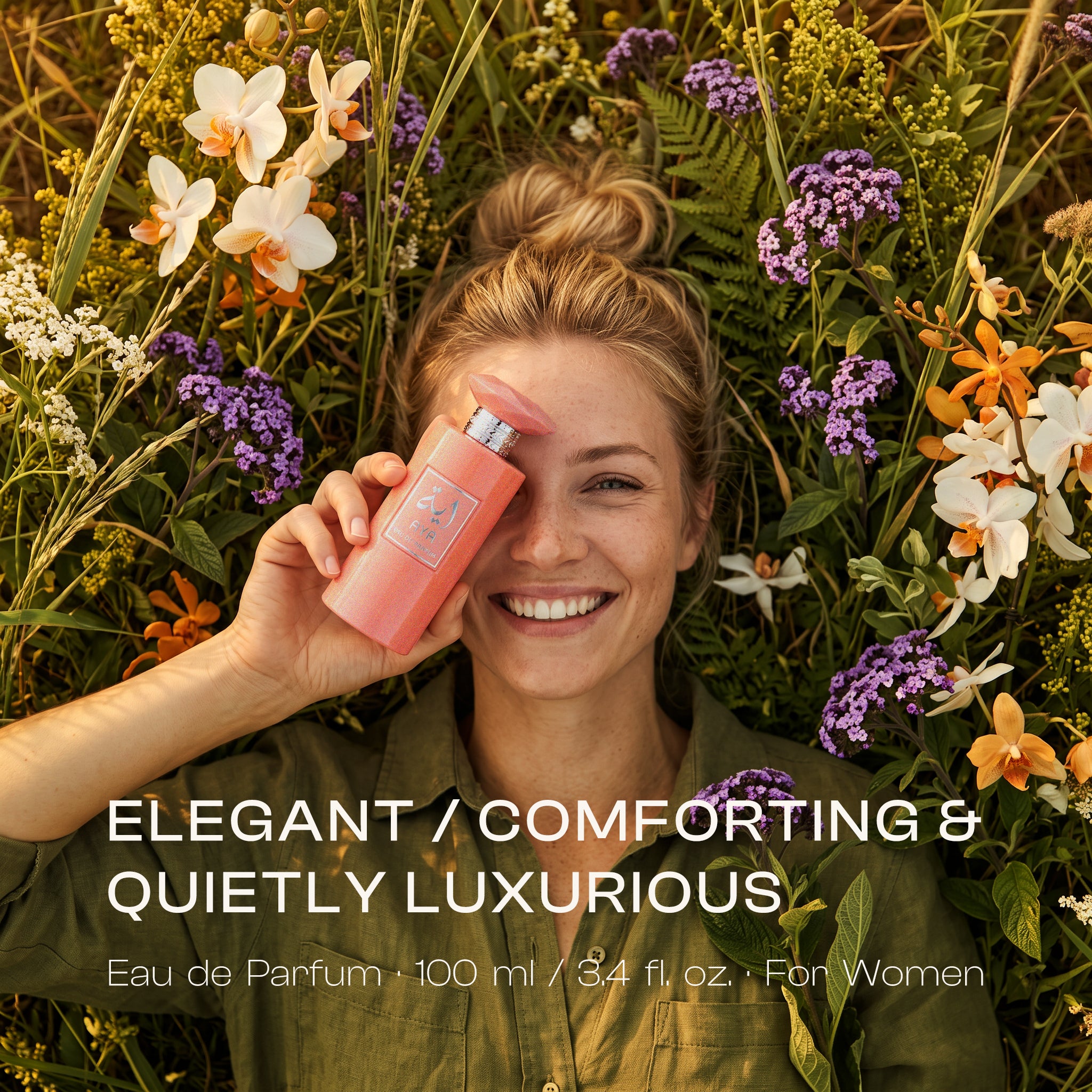 Smiling blonde woman lying in a field surrounded by white, orange, and purple flowers, holding a shimmery coral pink AYA Eau de Parfum bottle over one eye, with text overlay reading "ELEGANT / COMFORTING & QUIETLY LUXURIOUS".
